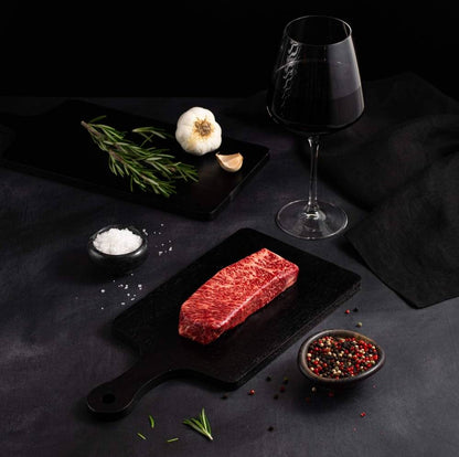 A Raw Australian Wagyu Full Blood Denver Steak rests on a black slate surrounded by salt, pepper, rosemary, garlic, and complemented by a glass of red wine on a dark surface.