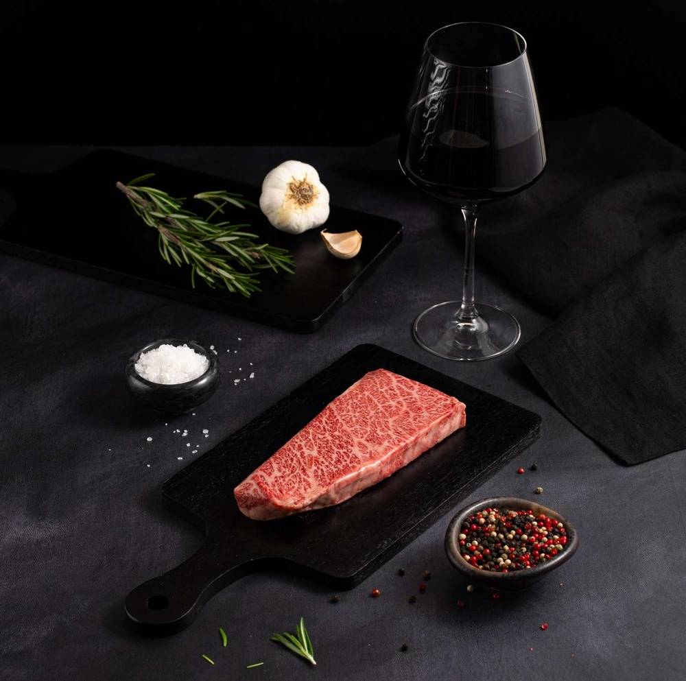 A Raw Japanese A5 Wagyu Denver Steak rests on a black slate surrounded by salt, pepper, rosemary, garlic, and complemented by a glass of red wine on a dark surface.