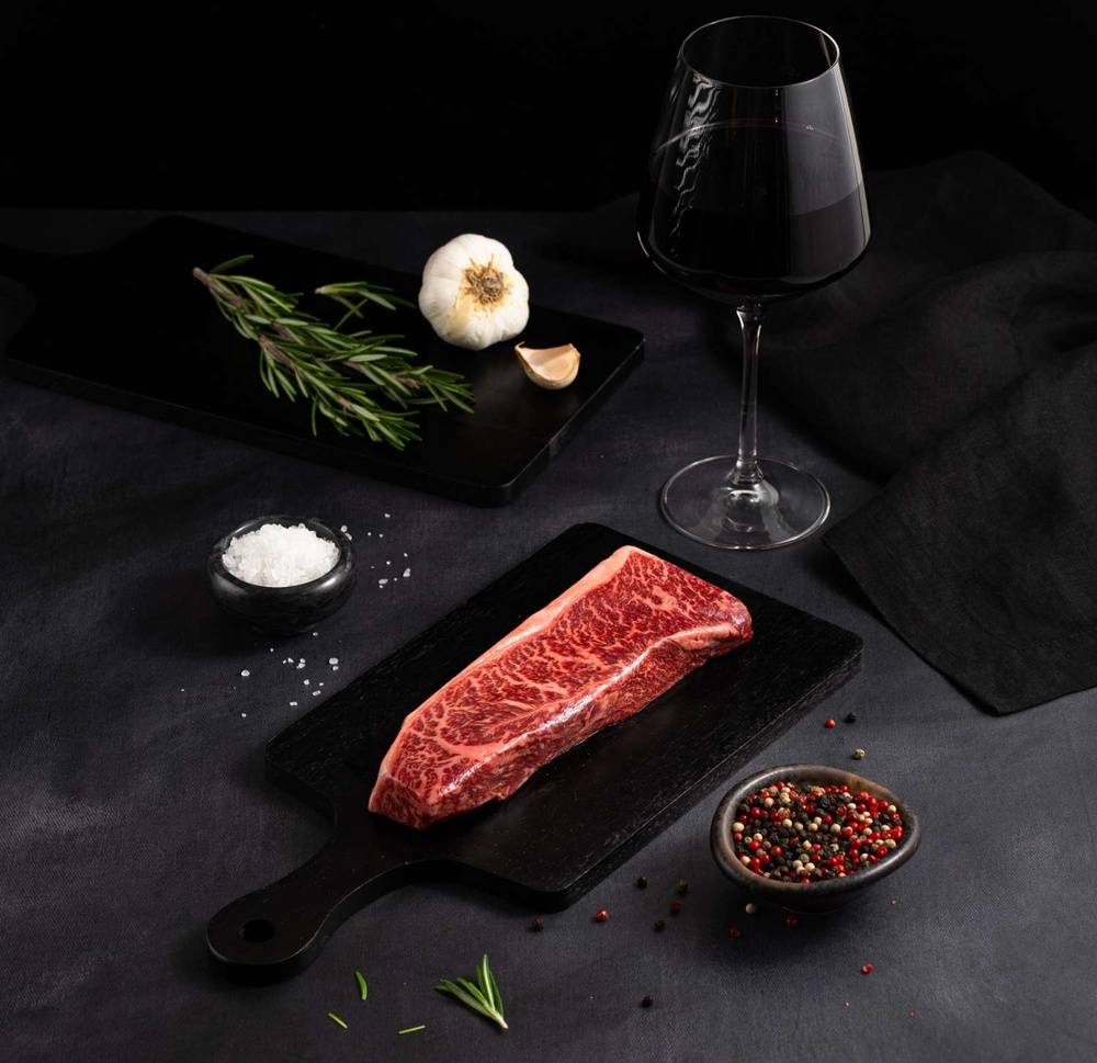 A Raw Australian Wagyu Top Coulotte Picanha Steak rests on a black slate surrounded by salt, pepper, rosemary, garlic, and complemented by a glass of red wine on a dark surface.