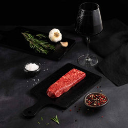 A Raw Australian Wagyu Denver Steak rests on a black slate surrounded by salt, pepper, rosemary, garlic, and complemented by a glass of red wine on a dark surface.