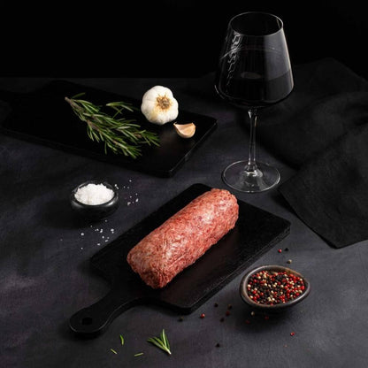 A slab of Japanese A5 Wagyu Ground Beef rests on a black slate surrounded by salt, pepper, rosemary, garlic, and complemented by a glass of red wine on a dark surface.