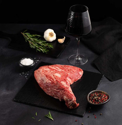 A succulent American Wagyu Masami Tri Tip, raw and marbled, rests on a black slate surrounded by salt, pepper, rosemary, garlic, and complemented by a glass of red wine on a dark surface.