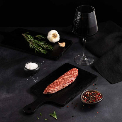 A Raw Japanese A5 Wagyu Flap Meat Steak rests on a black slate surrounded by salt, pepper, rosemary, garlic, and complemented by a glass of red wine on a dark surface.