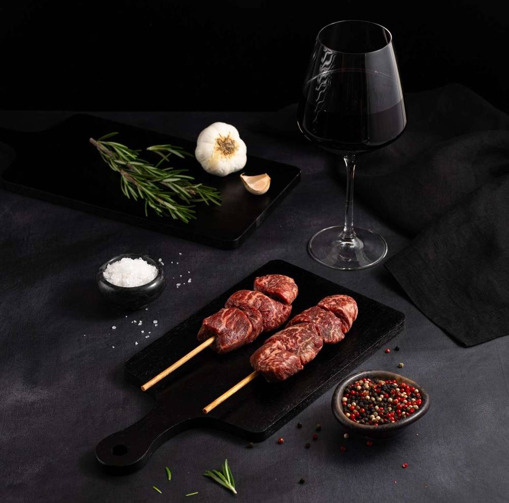 Two Australian Wagyu Filet Mignon Skewers rests on a black slate surrounded by salt, pepper, rosemary, garlic, and complemented by a glass of red wine on a dark surface.