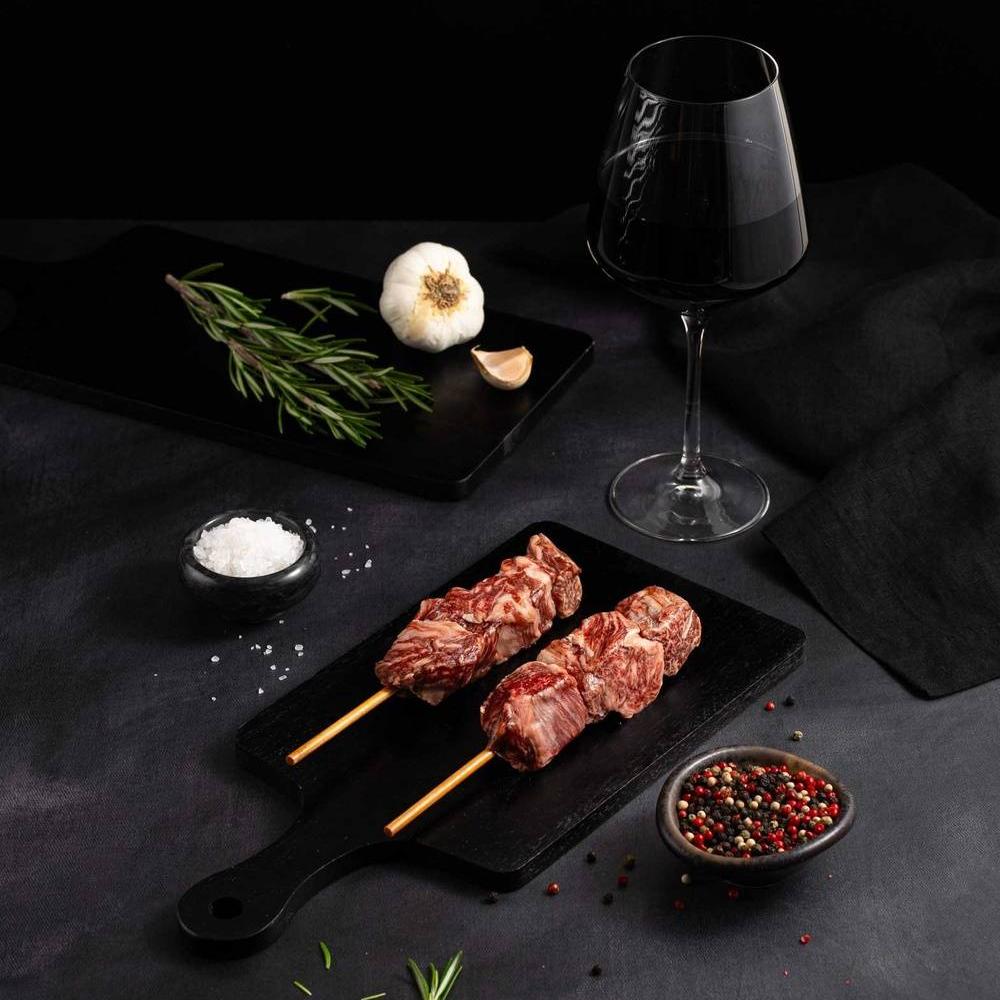 Two Raw Japanese A5 Wagyu Filet Mignon Skewers rests on a black slate surrounded by salt, pepper, rosemary, garlic, and complemented by a glass of red wine on a dark surface.