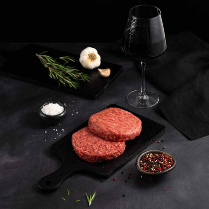 Two Australian Wagyu Beef Patties rests on a black slate surrounded by salt, pepper, rosemary, garlic, and complemented by a glass of red wine on a dark surface.