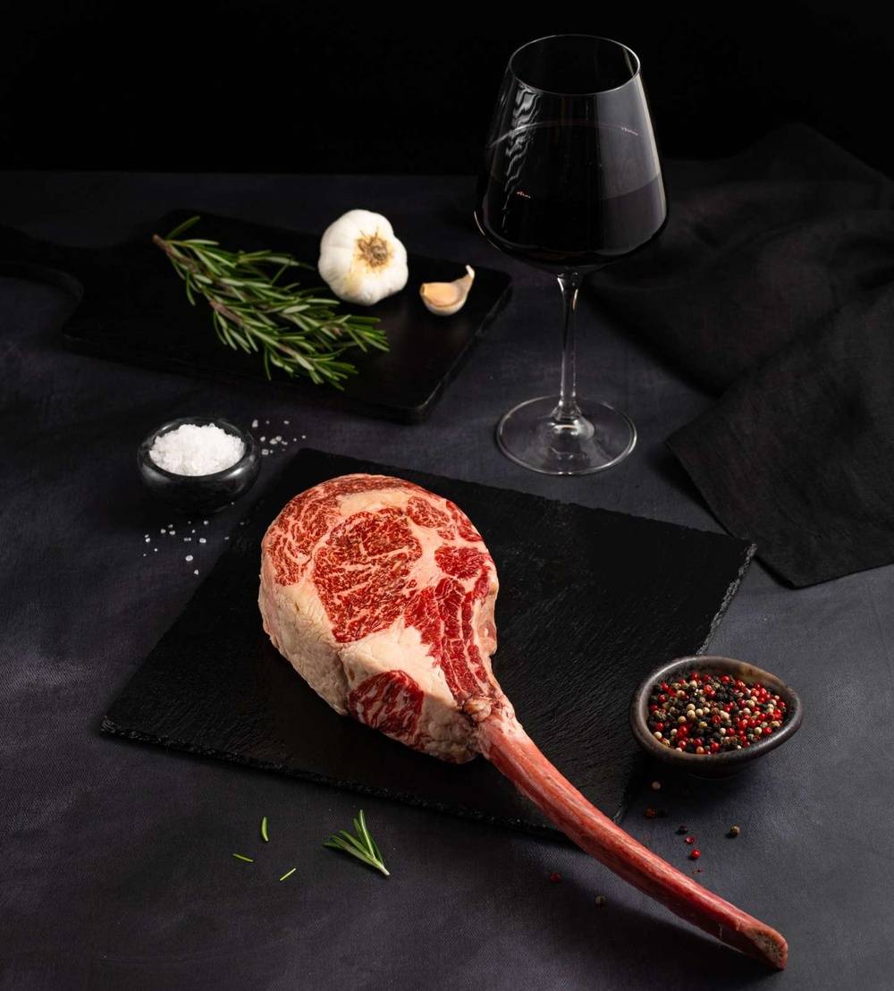 A Raw Australian Wagyu Tomahawk Steak rests on a black slate surrounded by salt, pepper, rosemary, garlic, and complemented by a glass of red wine on a dark surface.