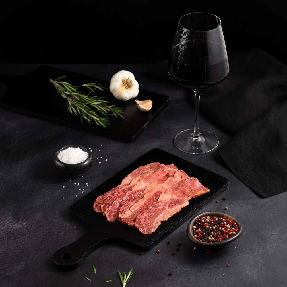 A Raw Australian Wagyu Beef Bacon rests on a black slate surrounded by salt, pepper, rosemary, garlic, and complemented by a glass of red wine on a dark surface.