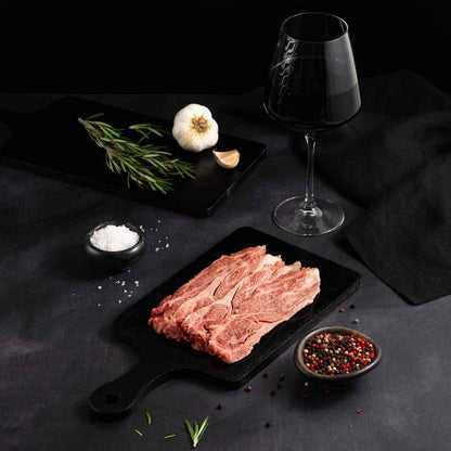 A Raw Japanese A5 Wagyu Sliced Chuck Roll rests on a black slate surrounded by salt, pepper, rosemary, garlic, and complemented by a glass of red wine on a dark surface.