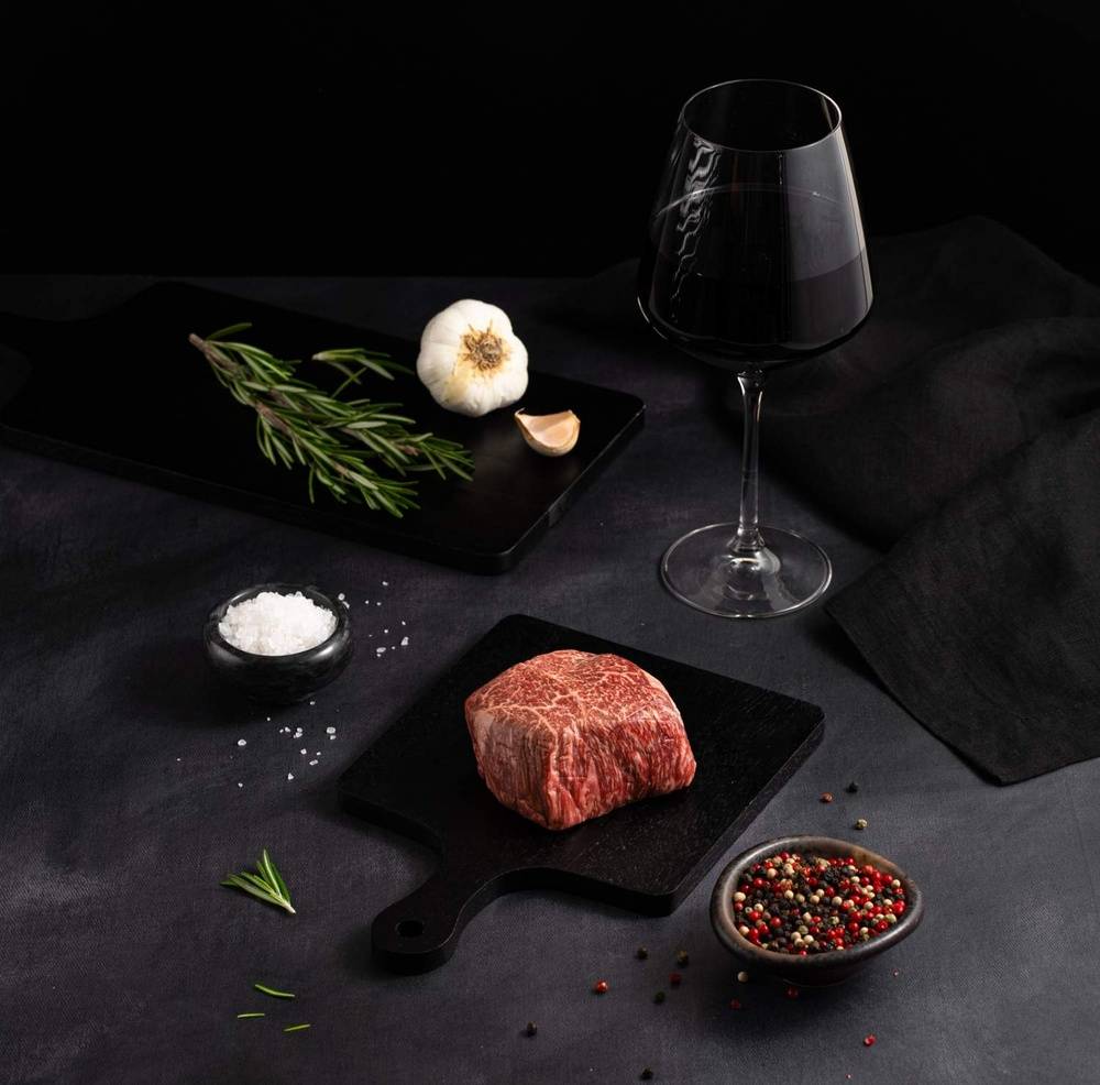 A Raw Japanese A5 Wagyu Top Sirloin rests on a black slate surrounded by salt, pepper, rosemary, garlic, and complemented by a glass of red wine on a dark surface.