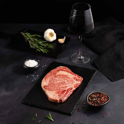 A Raw Japanese A5 Wagyu Miyazaki Ribeye Steak rests on a black slate surrounded by salt, pepper, rosemary, garlic, and complemented by a glass of red wine on a dark surface.