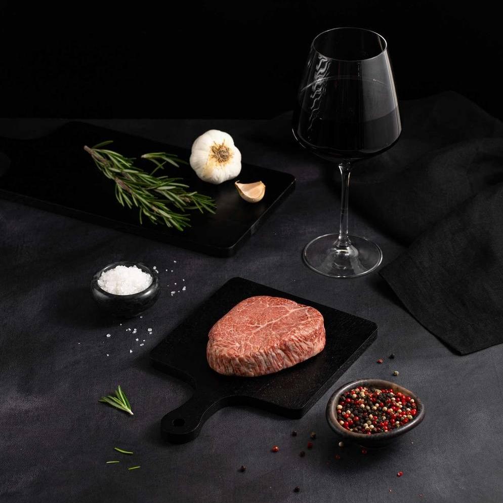 A Raw Japanese A5 Wagyu Kobe Wine Filet Steak rests on a black slate surrounded by salt, pepper, rosemary, garlic, and complemented by a glass of red wine on a dark surface.