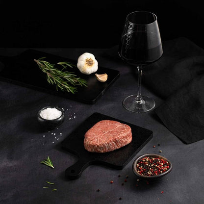 A Raw Japanese A5 Wagyu Kobe Wine Filet Steak rests on a black slate surrounded by salt, pepper, rosemary, garlic, and complemented by a glass of red wine on a dark surface.