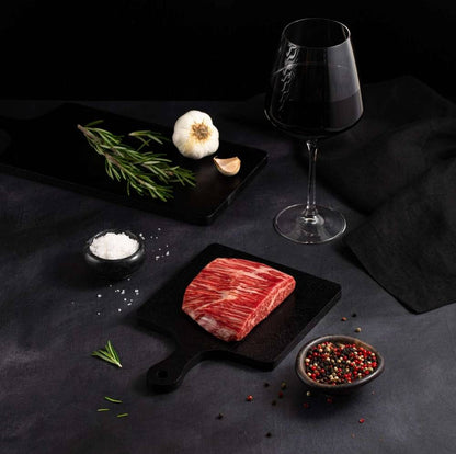 A Raw Australian Wagyu Flank Steak rests on a black slate surrounded by salt, pepper, rosemary, garlic, and complemented by a glass of red wine on a dark surface.