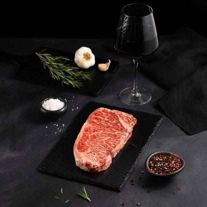 A Raw Japanese A5 Wagyu Kobe Wine New York Strip Steak rests on a black slate surrounded by salt, pepper, rosemary, garlic, and complemented by a glass of red wine on a dark surface.