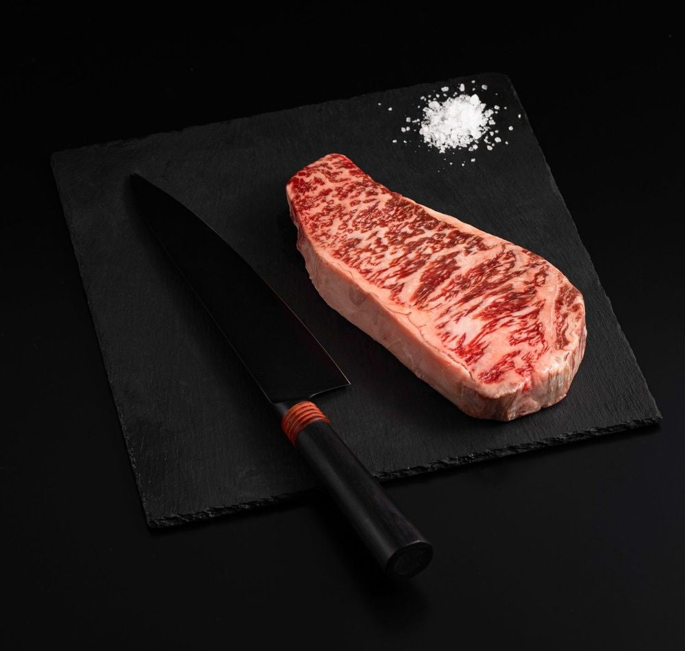 An Australian Wagyu Full Blood New York Striploin Steak with salt and chef's knife on a black slate, isolated on black background.