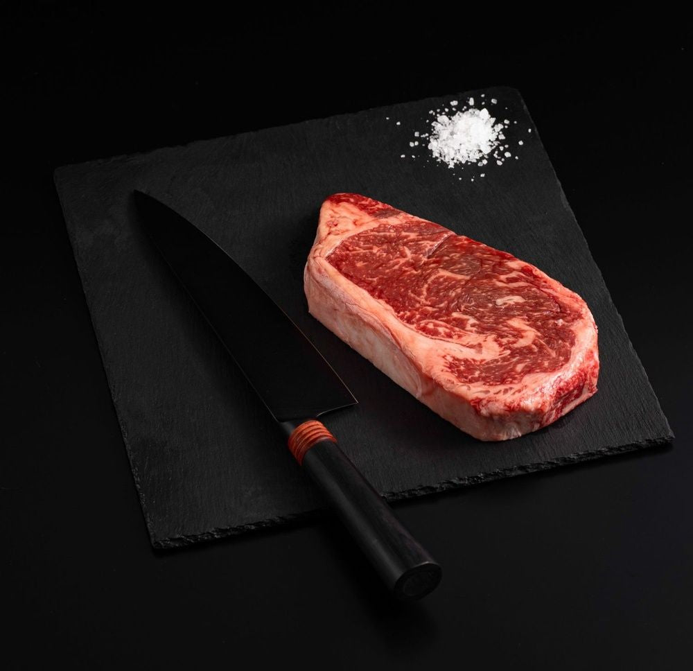 Raw marbled American Wagyu New York Masami Steak with salt and chef's knife on a black slate, isolated on black background.