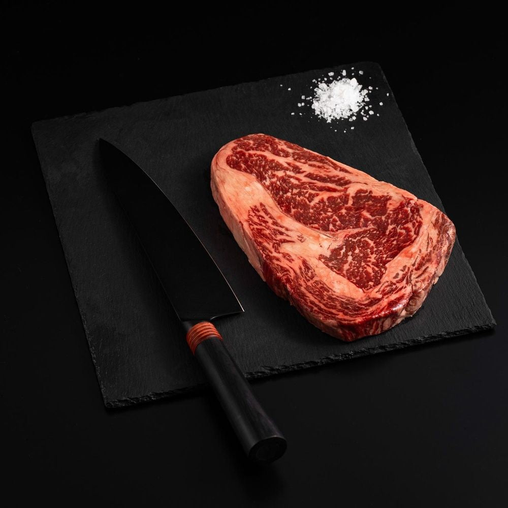 An Australian Wagyu Jade Full Blood Ribeye Steak with salt and chef's knife on a black slate, isolated on black background.