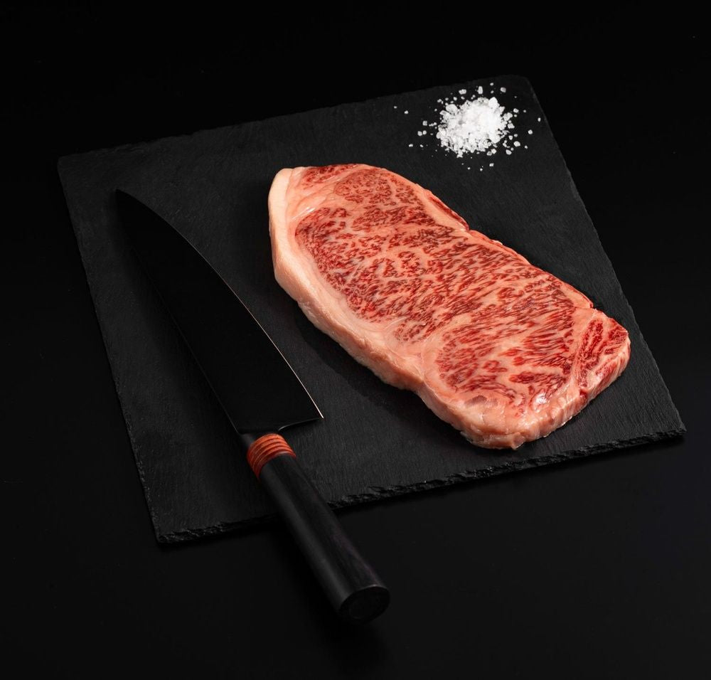 A Japanese A5 Wagyu Kobe Wine New York Strip Steak with salt and chef's knife on a black slate, isolated on black background.