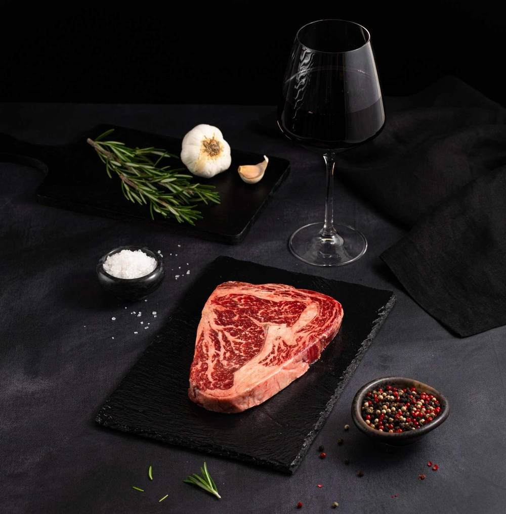 A Raw Australian Wagyu Jade Full Blood Ribeye Steak rests on a black slate surrounded by salt, pepper, rosemary, garlic, and complemented by a glass of red wine on a dark surface.