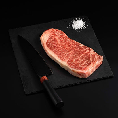 A Japanese A5 Wagyu Striploin Miyazaki Steak with salt and chef's knife on a black slate, isolated on black background.