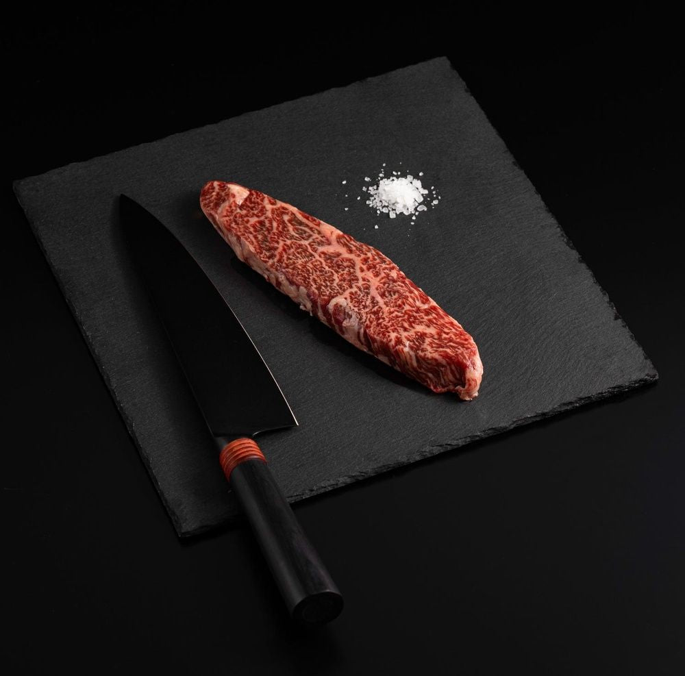 A Japanese A5 Wagyu Flap Meat Steak with salt and chef's knife on a black slate, isolated on black background.