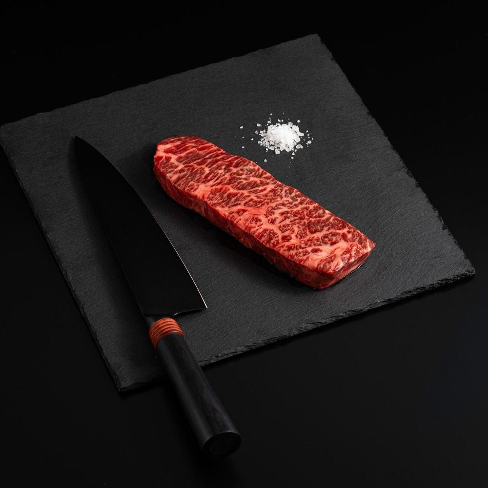 An Australian Wagyu Denver Steak with salt and chef's knife on a black slate, isolated on black background.