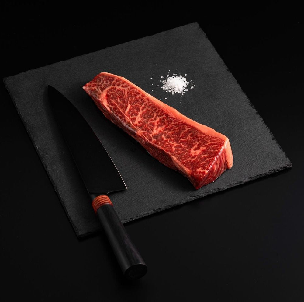 An Australian Wagyu Top Coulotte Picanha Steak with salt and chef's knife on a black slate, isolated on black background.