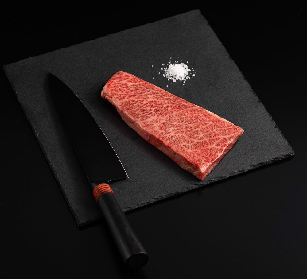 A Japanese A5 Wagyu Denver Steak with salt and chef's knife on a black slate, isolated on black background.