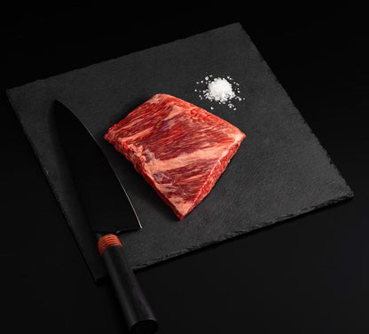 A Japanese A5 Wagyu Rib Cap Steak with salt and chef's knife on a black slate, isolated on black background.