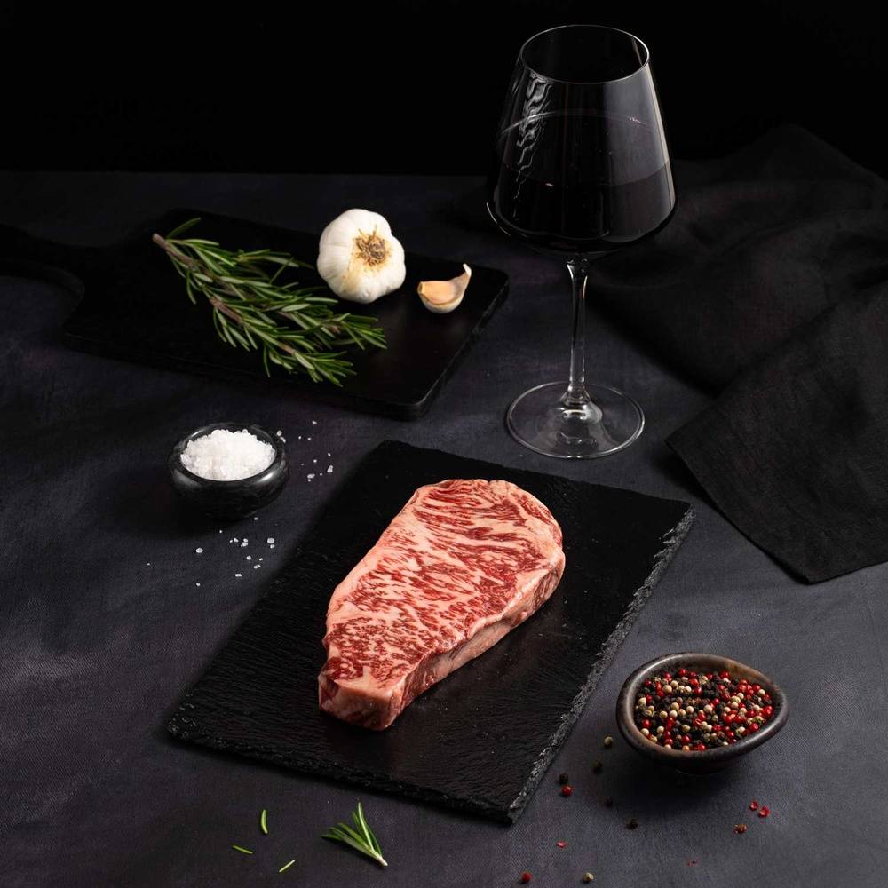 A Raw Australian Wagyu Full Blood New York Striploin Steak rests on a black slate surrounded by salt, pepper, rosemary, garlic, and complemented by a glass of red wine on a dark surface.