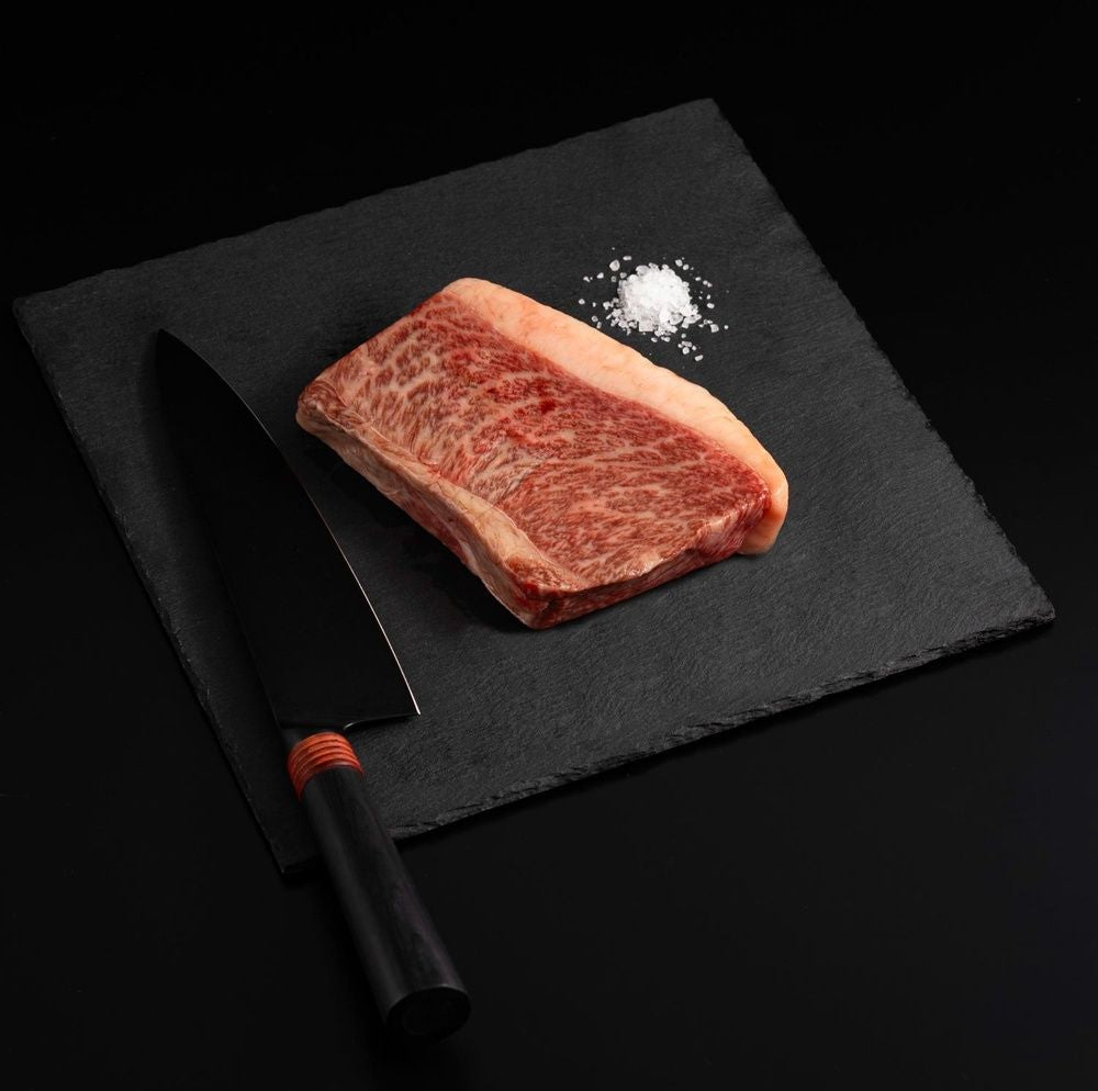 A Japanese A5 Top Coulotte Wagyu Picanha Steak with salt and chef's knife on a black slate, isolated on black background.