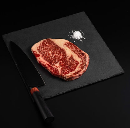 An Australian Wagyu Tomahawk Ribeye Steak with salt and chef's knife on a black slate, isolated on black background.