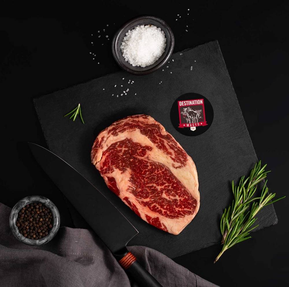 An Australian Wagyu Tomahawk Ribeye Steak rests on a slate board with rosemary, a bowl of salt, a bowl of pepper, and a black knife.