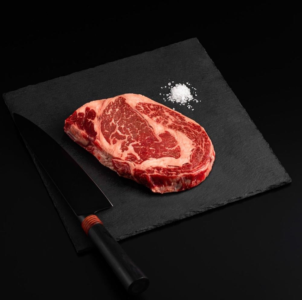 Raw marbled American Wagyu Masami Ribeye Steak with salt and chef's knife on a black slate, isolated on black background.