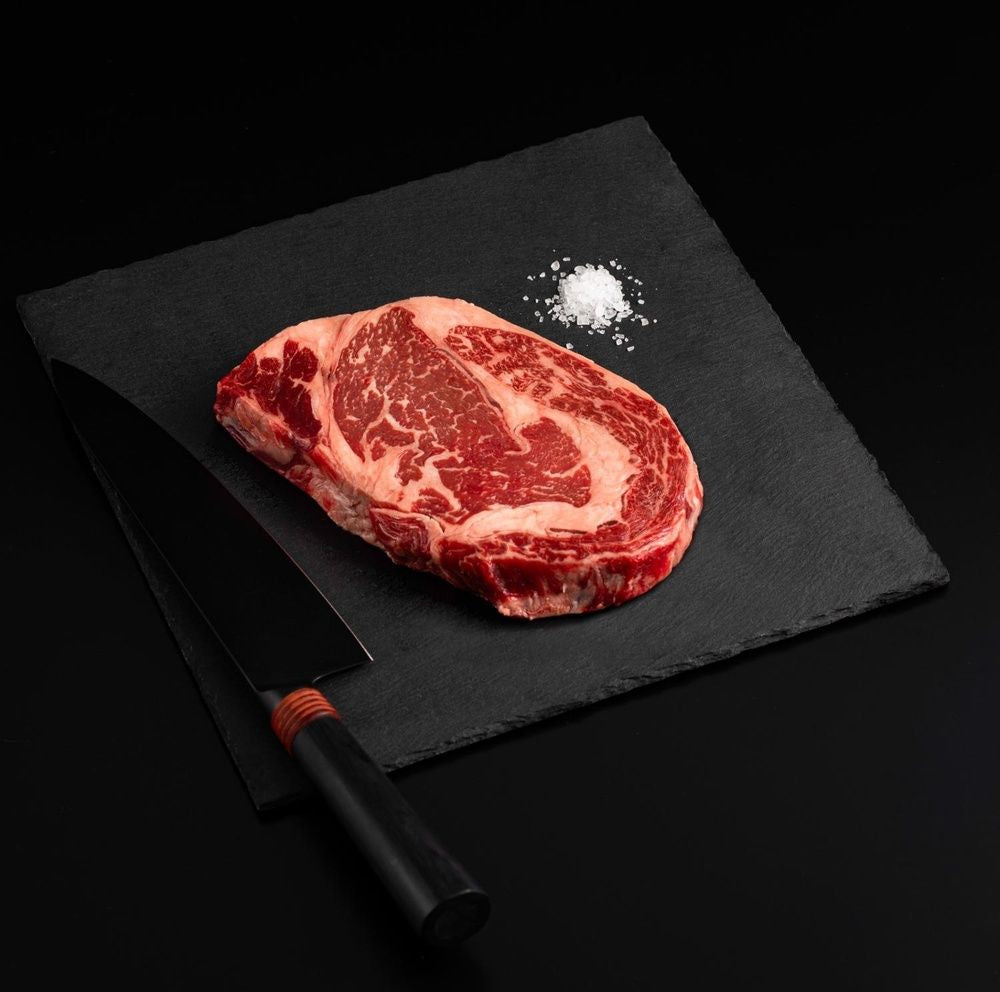 An Australian Wagyu Ribeye Steak with salt and chef's knife on a black slate, isolated on black background.