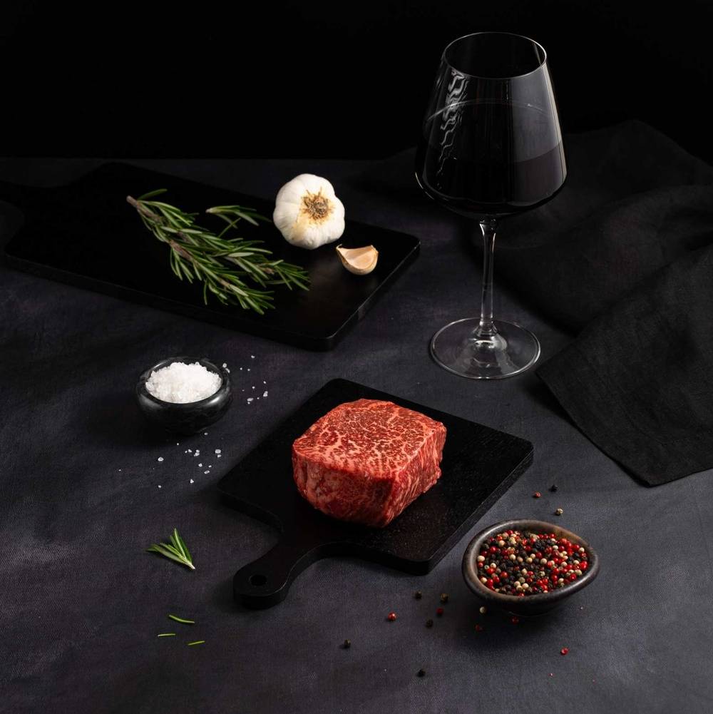 A Raw Australian Wagyu Full Blood Top Sirloin Steak rests on a black slate surrounded by salt, pepper, rosemary, garlic, and complemented by a glass of red wine on a dark surface.