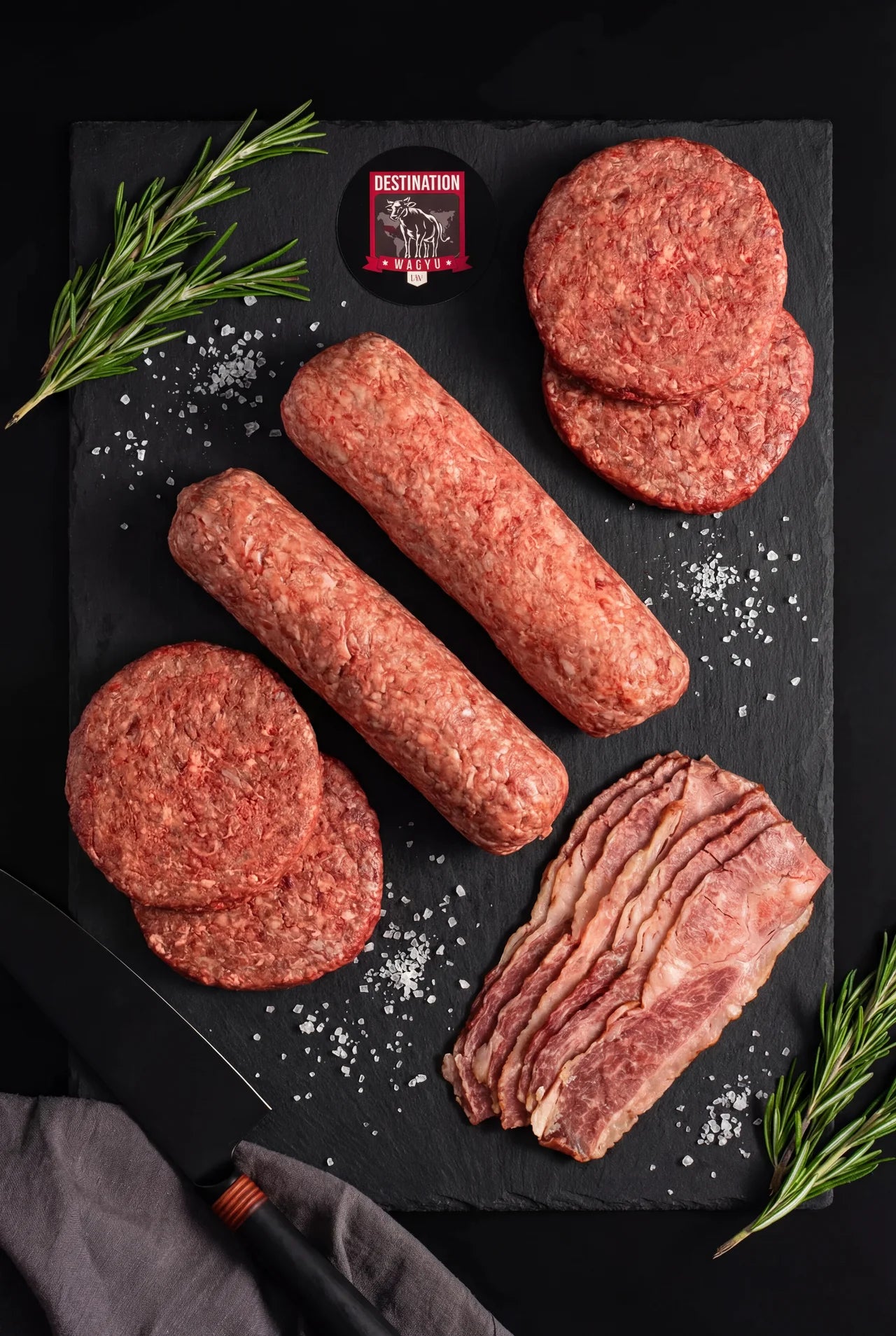 The Wagyu Burger Experience Box — four Australian Wagyu burger patties, two Japanese A5 Wagyu ground beef logs, and wood-smoked Australian Wagyu beef bacon on a black slate board with rosemary and sea salt, by Destination Wagyu.
