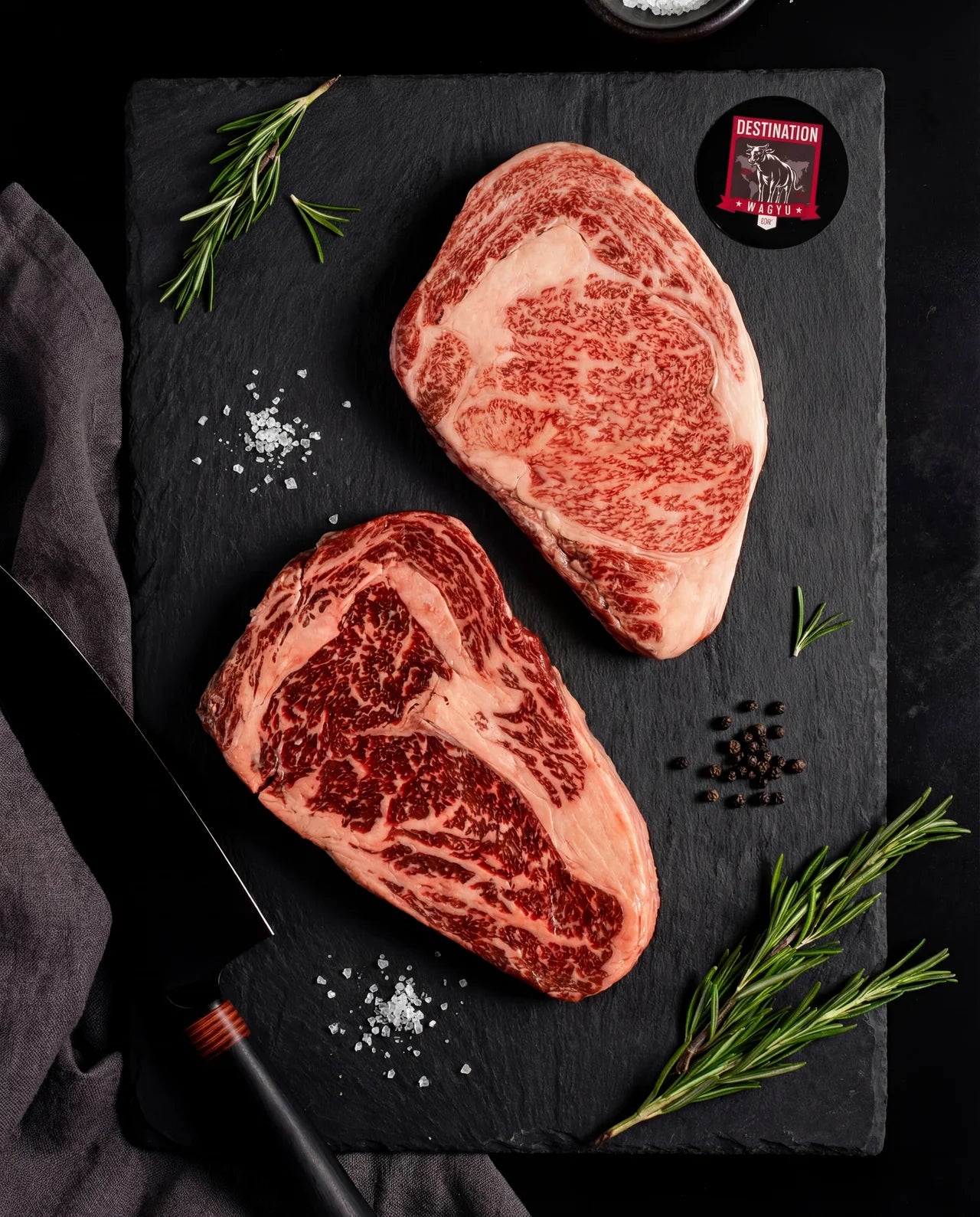 An Australian wagyu stone axe full blood ribeye steak with a Japanese a5 Kagoshima ribeye steak. Black board with black knife. Salt and pepper garnish