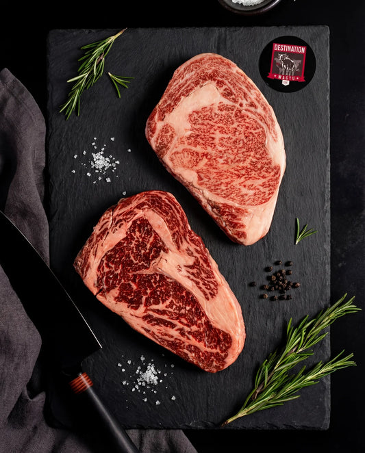 An Australian wagyu stone axe full blood ribeye steak with a Japanese a5 Kagoshima ribeye steak. Black board with black knife. Salt and pepper garnish